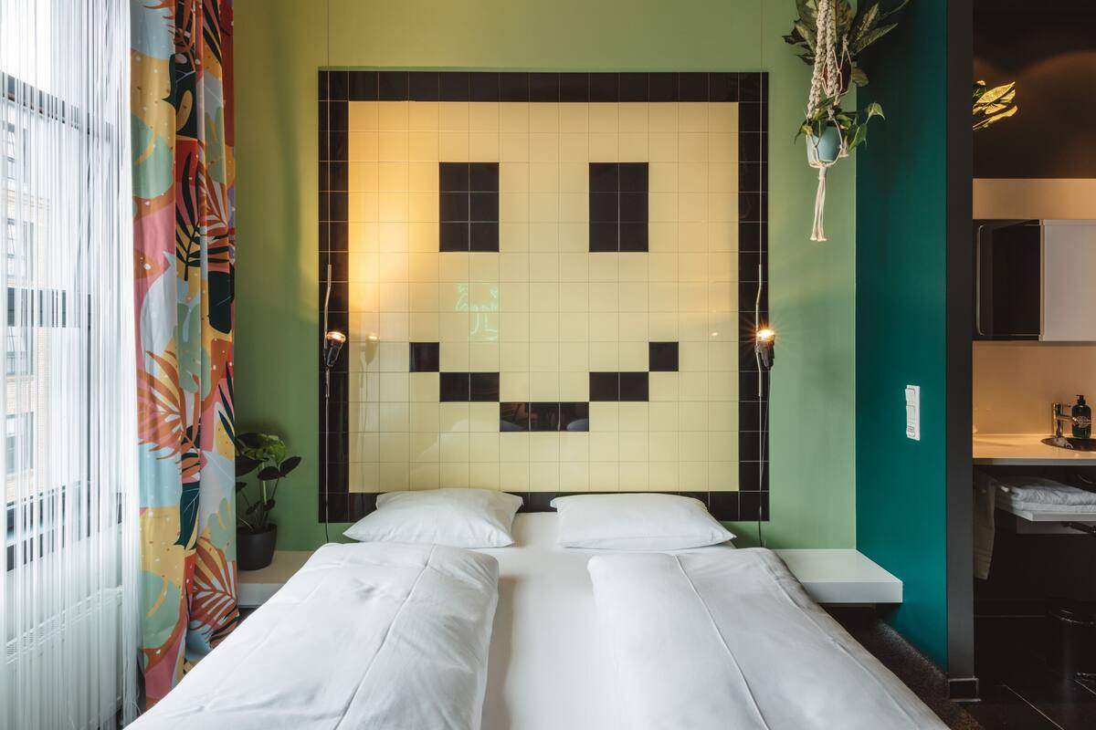 A hotel room with a bed, a small bedside table, a potted plant, and a wall with a geometric pattern.