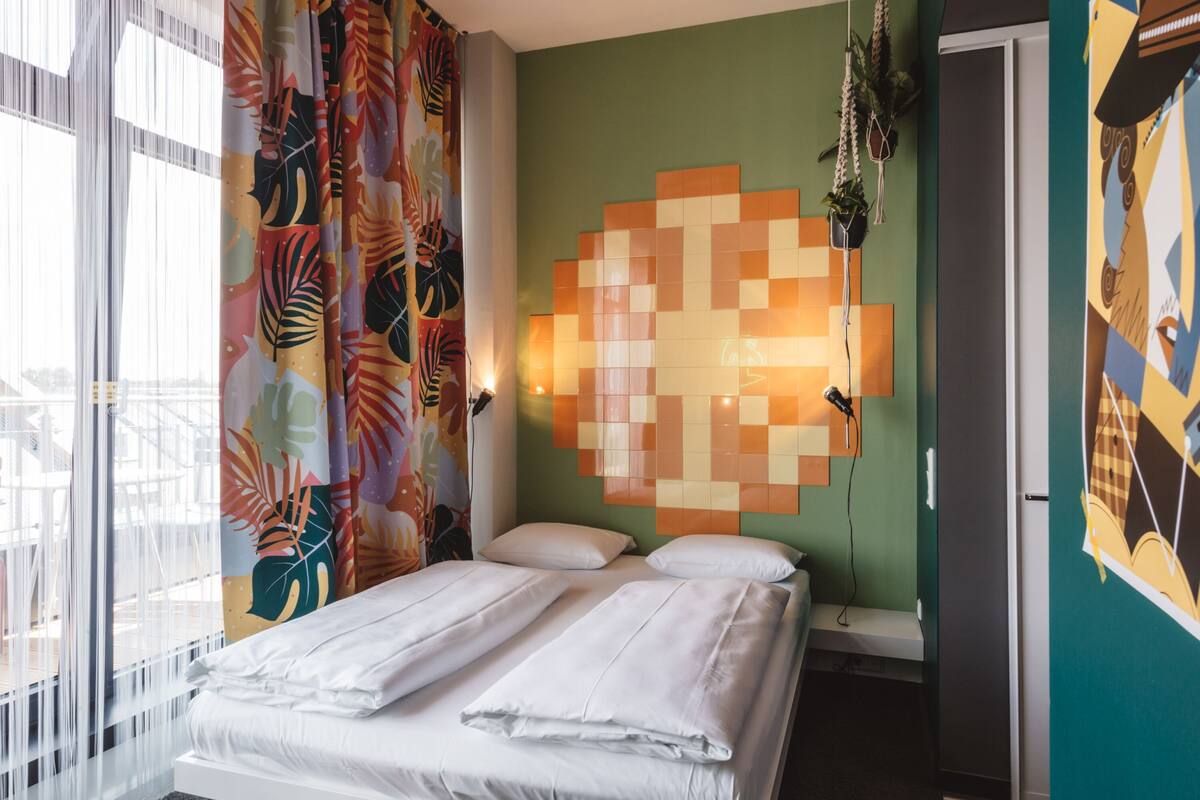 A hotel room with a bed, curtains, a wall-mounted light, and a door.