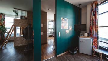 A modern living space with a wooden floor, a teal accent wall, a small kitchenette, and a view through a large window.