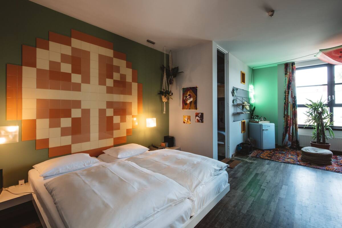 A modern bedroom with a large bed, a wall-mounted pixel art headboard, bedside lamps, a small desk, and a view of the hallway and balcony.