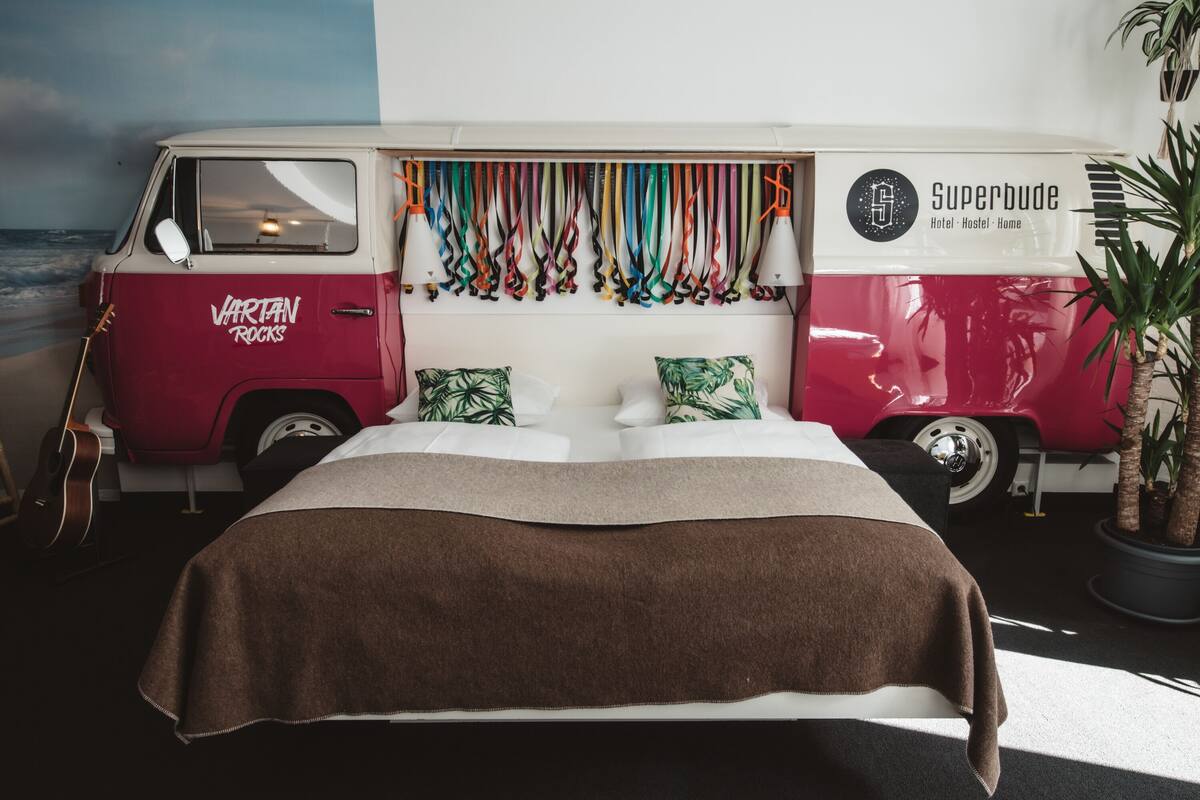A bed with a brown blanket, a red and white van with 'Vartan Rocks' and 'Superbude' branding, and a guitar leaning against the van.