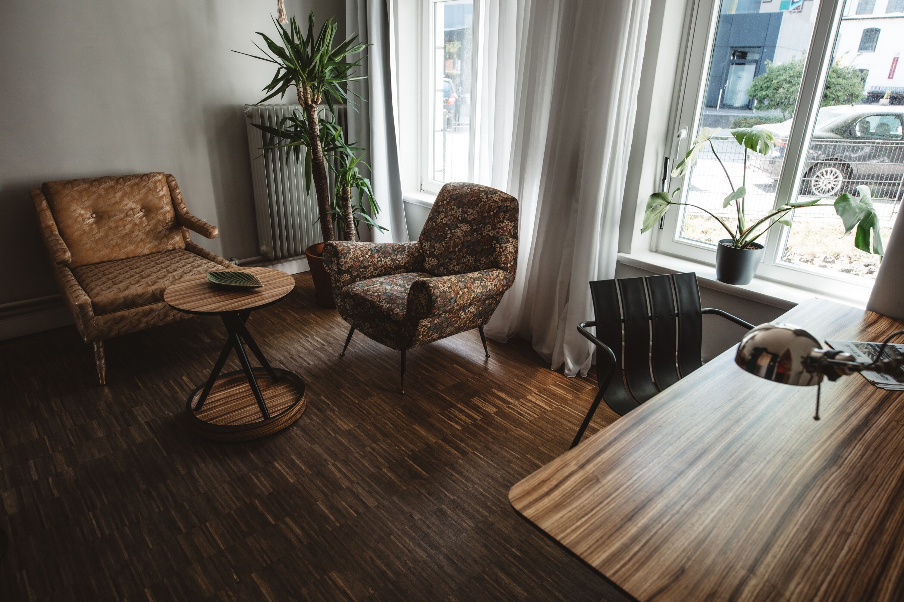 Un salon moderne avec un plancher en bois, deux fauteuils, une petite table ronde, une plante en pot et une vue sur la rue par une fenêtre.