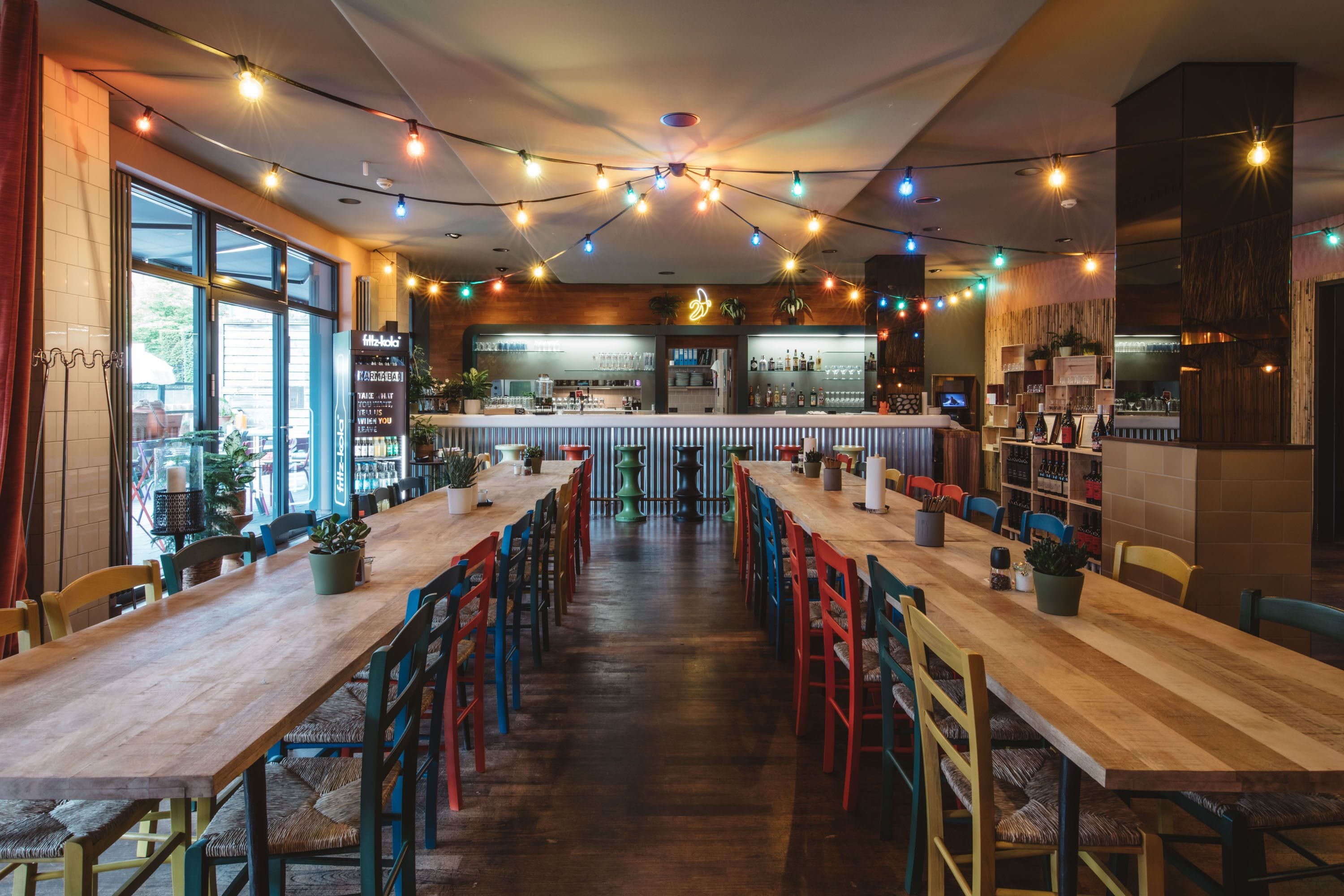 A restaurant with long wooden tables and colorful chairs, a bar area, and potted plants.