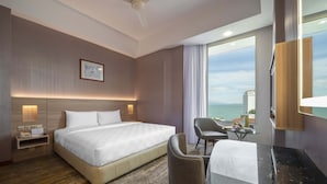 Habitación de hotel con una cama grande, un escritorio con silla, una mesita, una mesita de noche, un ventilador de techo y un ventanal con vista a un paisaje urbano.