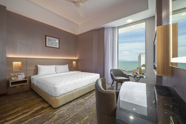 A hotel room with a large bed, a desk with a chair, a small table, a nightstand, a ceiling fan, and a large window with a view of a cityscape. - Cititel Penang (George Town)
