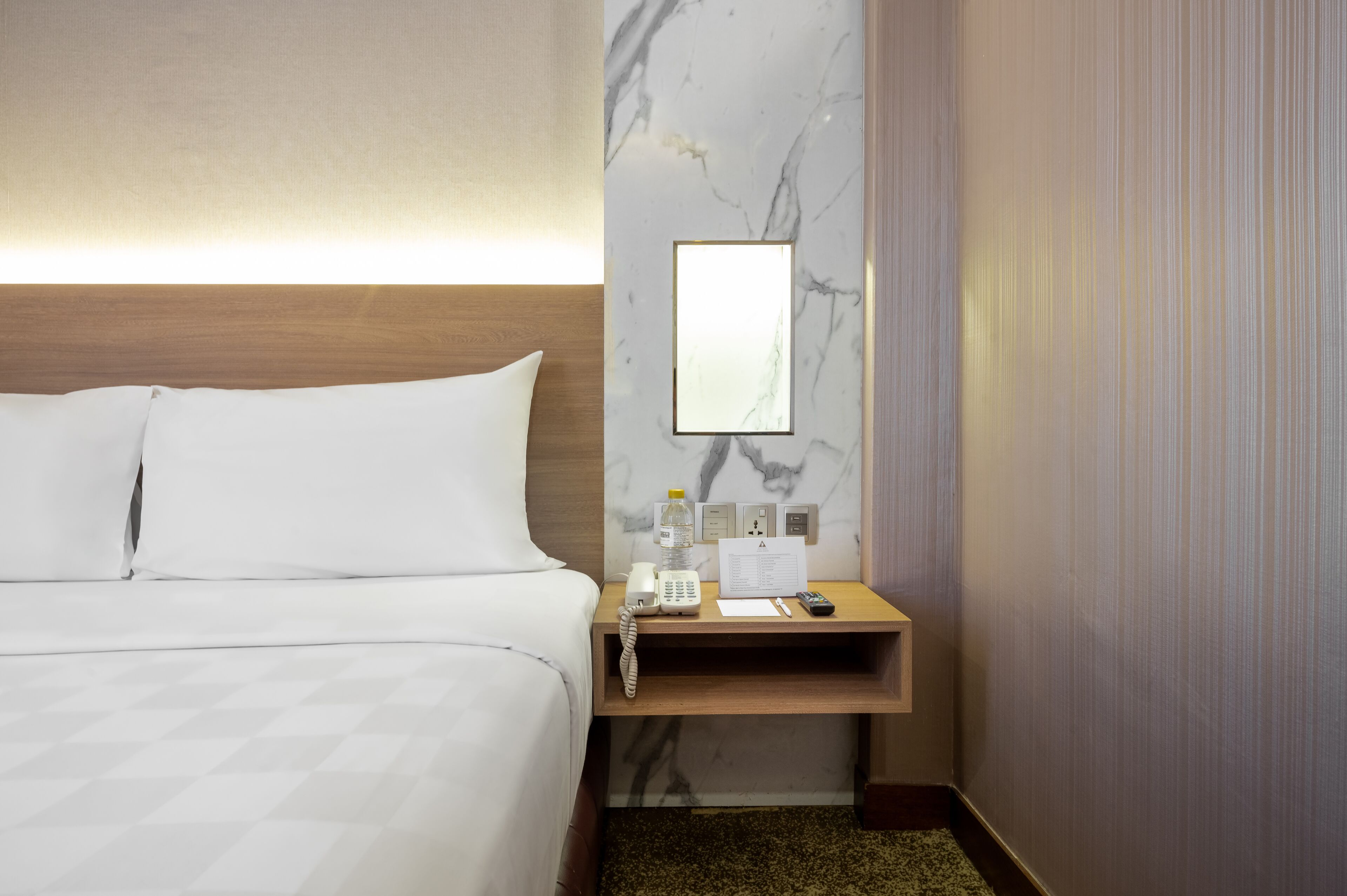 A hotel room with a bed, a wooden nightstand, a marble wall, a mirror, a phone, a water bottle, a notepad, and a remote control.