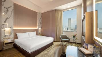A hotel room with a large bed, a desk with a chair, a small table with a plant, and a city view through the window.