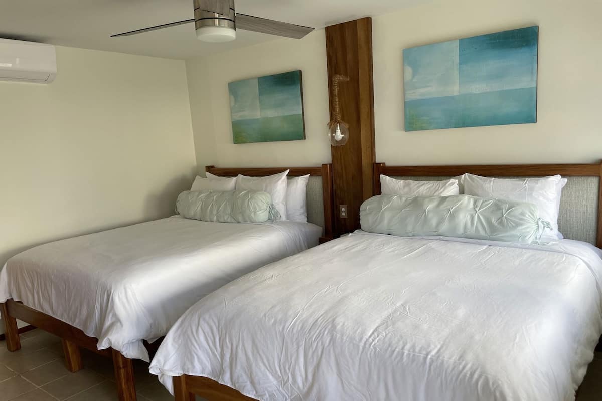 Habitación de hotel con dos camas, cabeceros de madera, ropa de cama blanca, ventilador de techo y dos cuadros colgados en la pared.