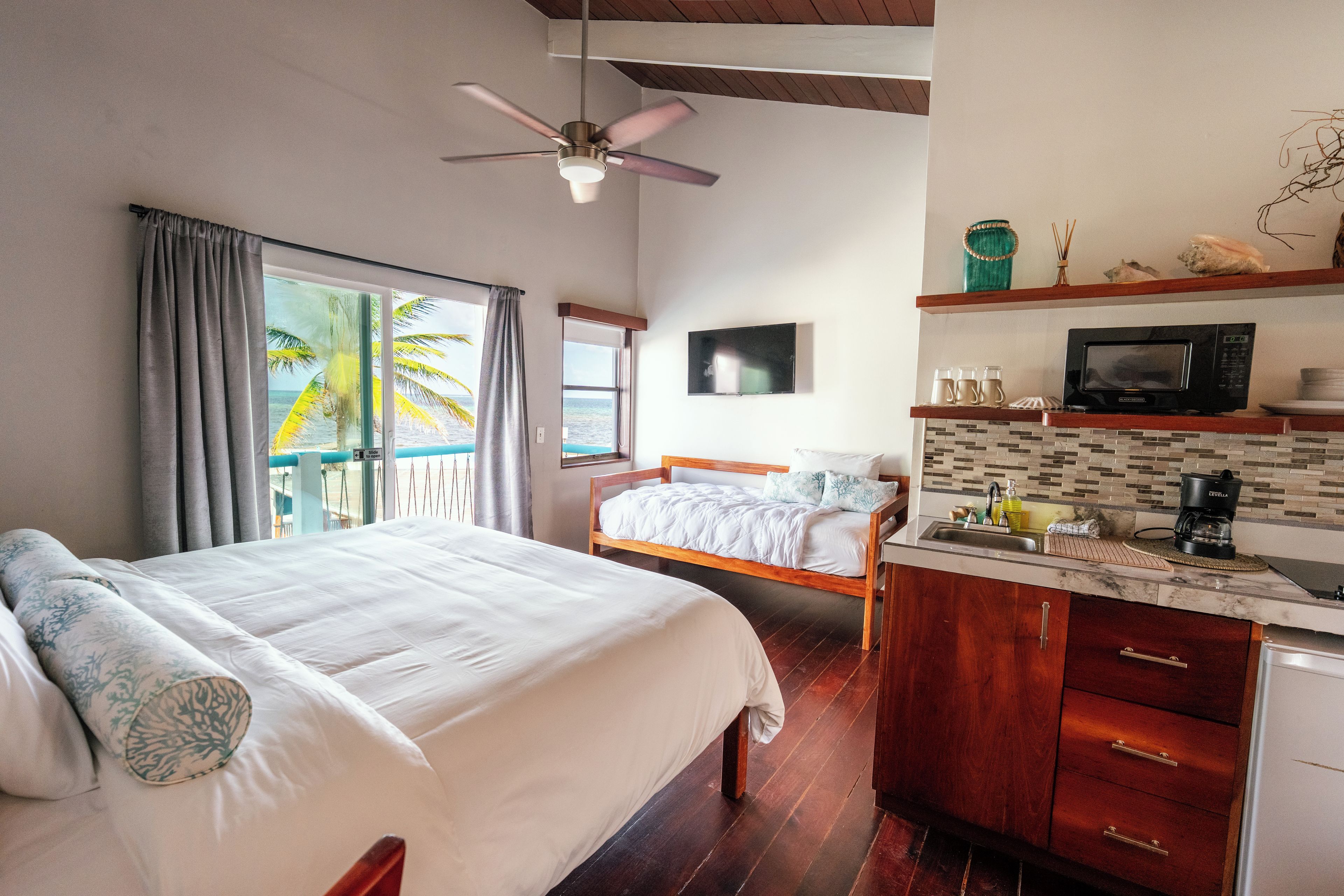 A hotel room with a bed, a desk, a kitchenette, and a view of the ocean.