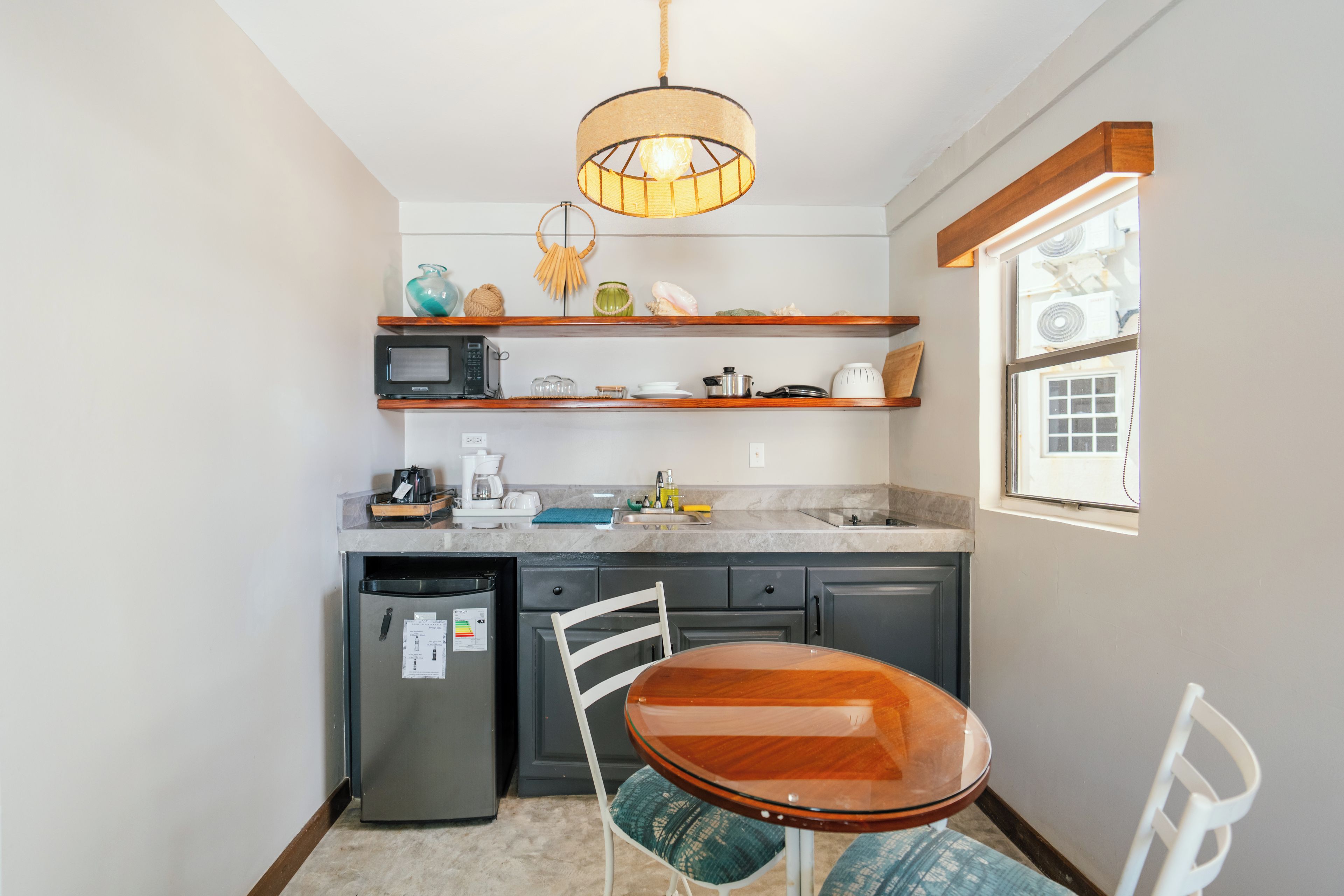 A compact kitchen with a built-in microwave, a coffee maker, and a small refrigerator. There is a wooden chair and a table with a glass top.