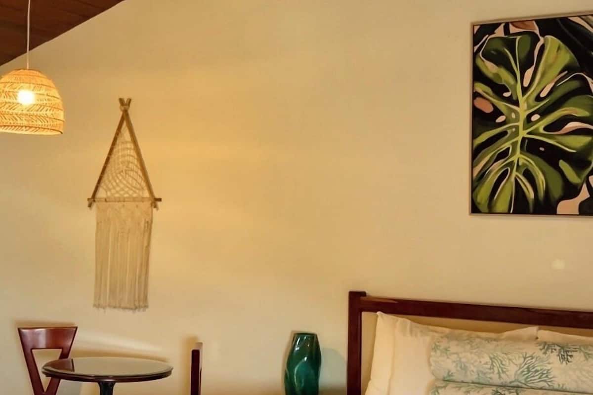 Un dormitorio con cama, ventilador de techo, lámpara colgante, silla, mesita pequeña y una planta sujeta a la pared.