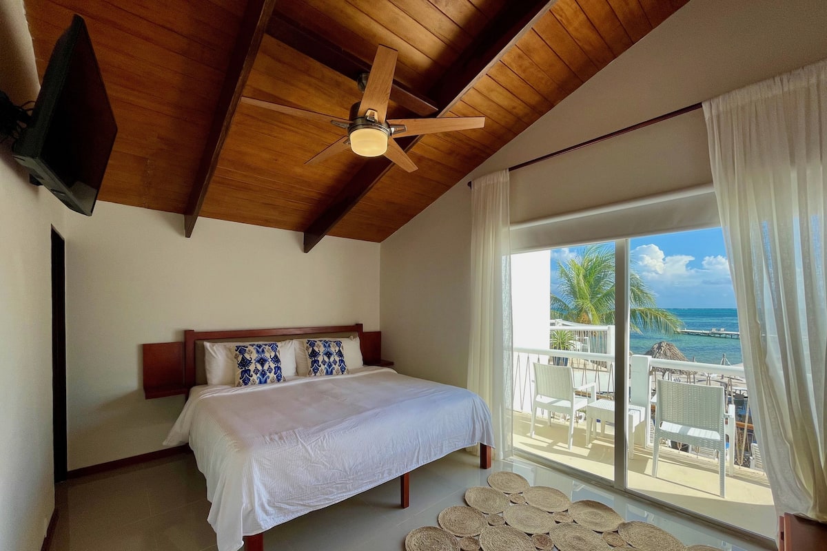 Un dormitorio con cama, ventilador de techo, televisión y balcón con vista al océano.