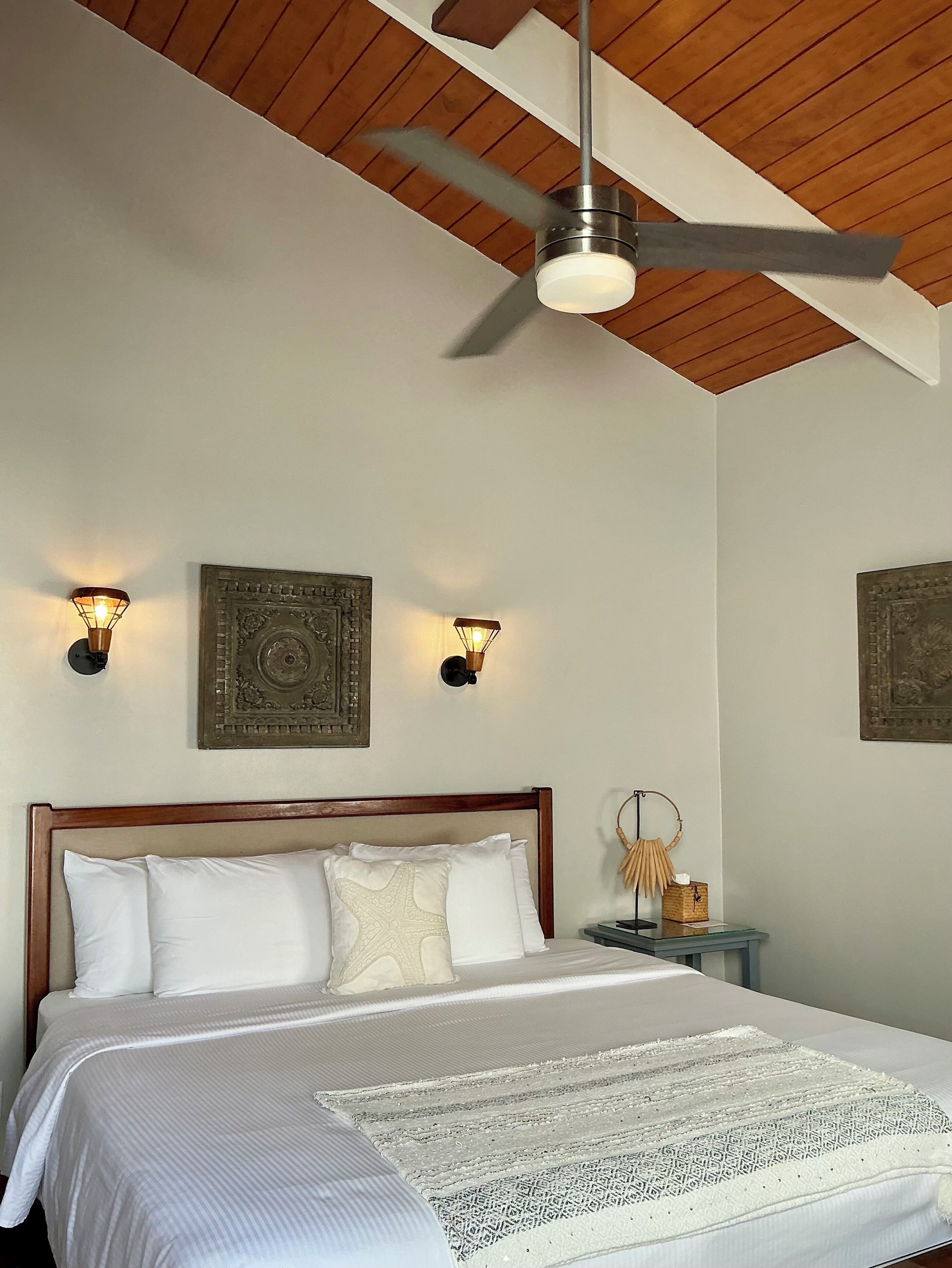 A bedroom with a wooden ceiling, a bed with white bedding, two wall-mounted light fixtures, and two decorative wall pieces.