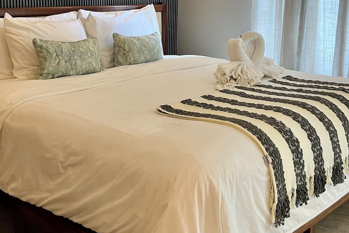 Una cama bien tendida con sábanas blancas, cabecera con rayas verticales, una lámpara de pared y una ventana con cortinas.
