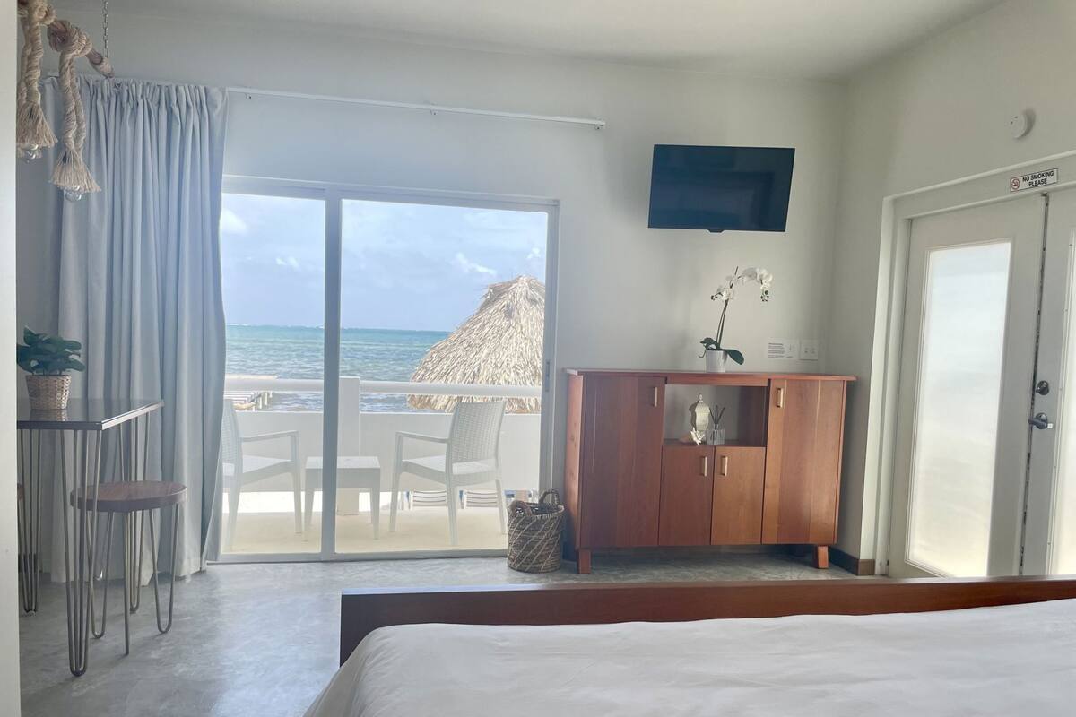 Una habitación de hotel con una cama, un televisor, un armario de madera, un balcón con vistas al océano y una sombrilla de paja.
