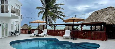 A swimming pool with lounge chairs and umbrellas, a thatched-roof structure, and a view of the ocean.