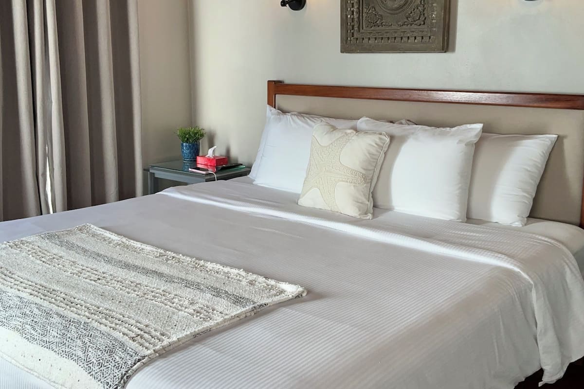 Una cama bien hecha con sábanas blancas y un cubrecolchón decorativo, una mesita de noche con lámpara, y un adorno en la pared.