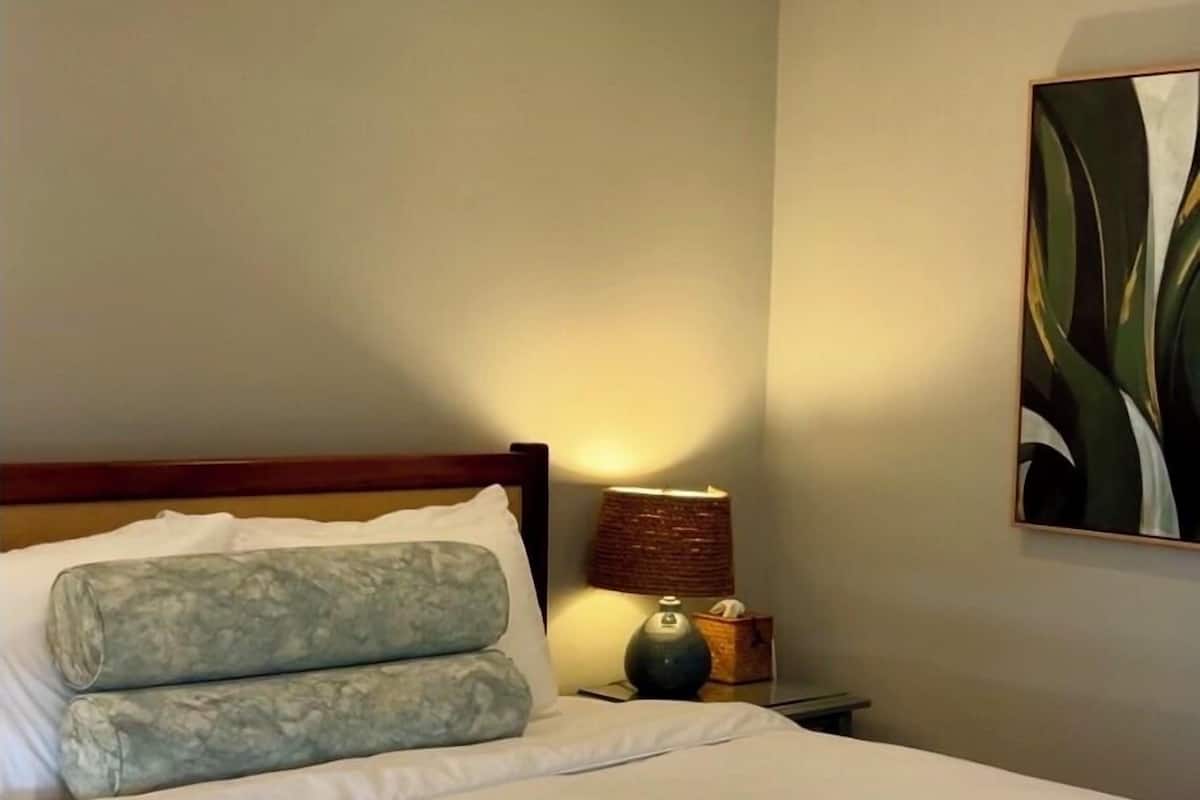 Habitación de hotel con cama, ventilador de techo, mesita de noche con lámpara y un cuadro en la pared.