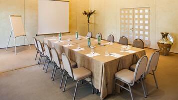 Un salón de conferencias con una larga mesa, sillas, una pantalla de proyector y un jarrón decorativo.
