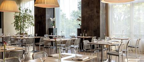 Un restaurant doté de nombreuses tables et chaises, de grands lustres suspendus et offrant une vue sur les arbres à travers les fenêtres.