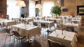 A spacious dining area with multiple tables and chairs, large windows with curtains, and a potted plant.