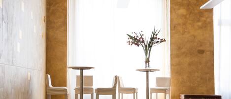 A room with a large window, two round tables, four chairs, a vase with flowers, and a vintage suitcase.
