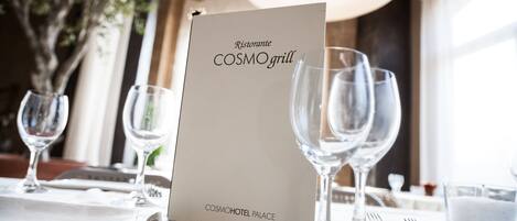 Una mesa dispuesta con copas de vino, cubiertos y una tarjeta del menú para Ristorante COSMOgrill, además de una vela sobre la mesa.