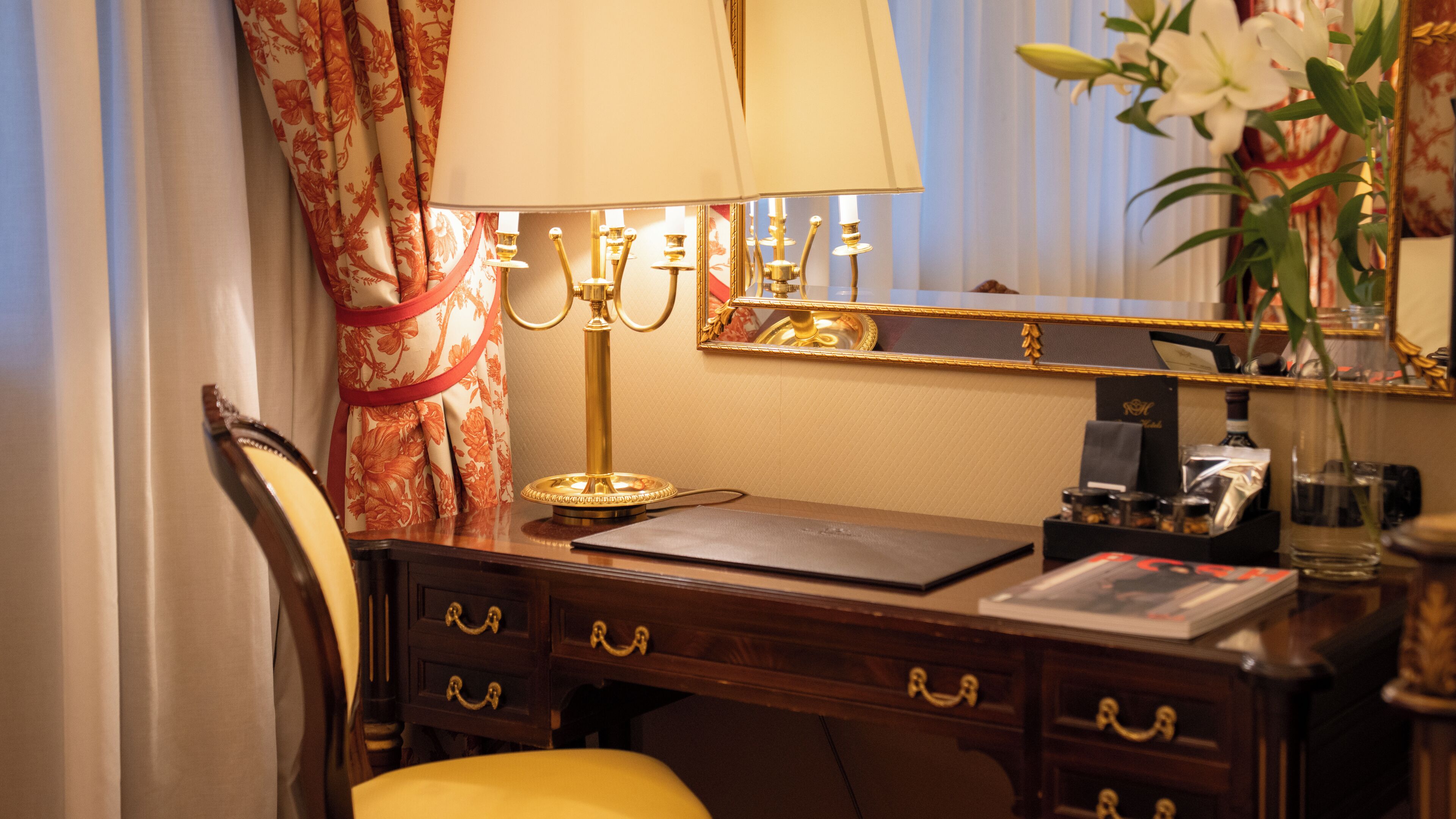 A wooden desk with a lamp, a mirror, and a vase with flowers.