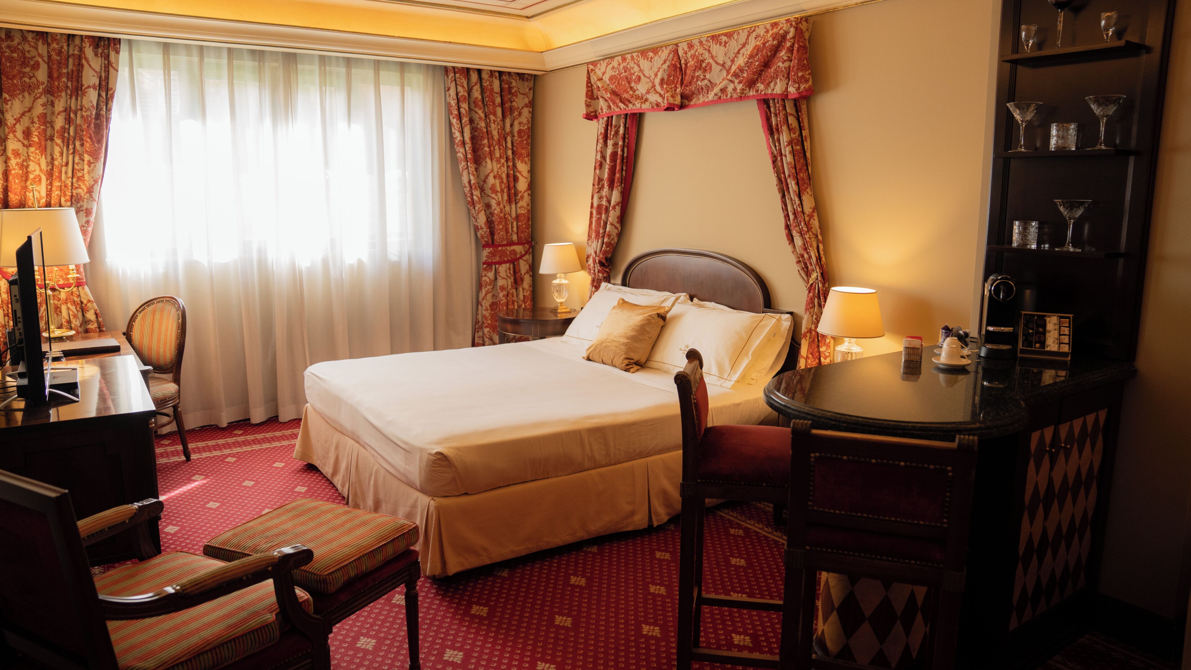 A hotel room with a bed, a desk, a chair, a table, and a shelf with glasses.