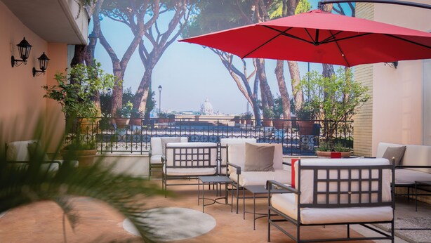 A terrace with outdoor seating, a red umbrella, and a view of trees and a distant building. - River Château Hotel (Rome)