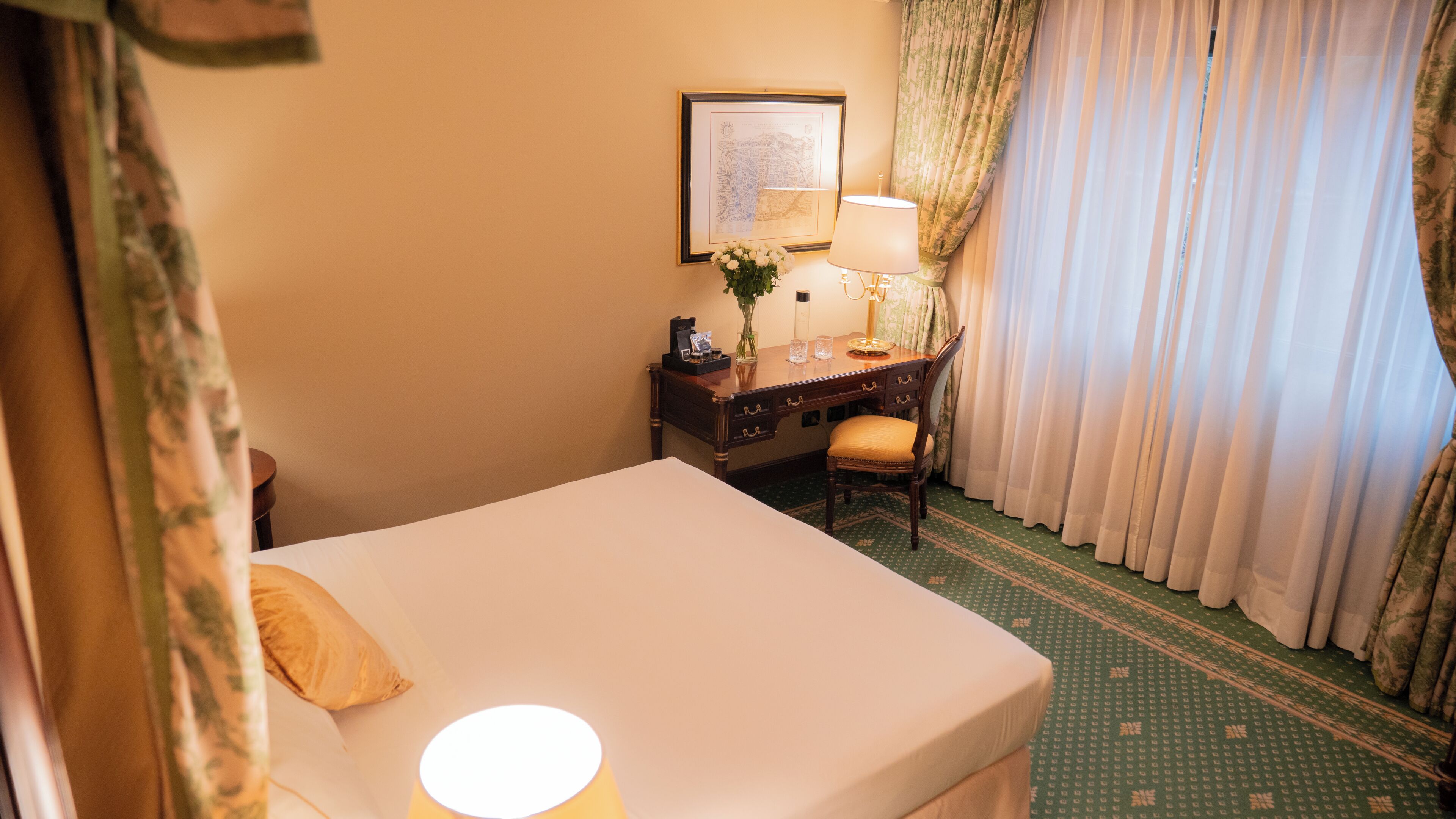 A hotel room with a bed, a desk with a lamp, a vase with flowers, and a framed map on the wall.