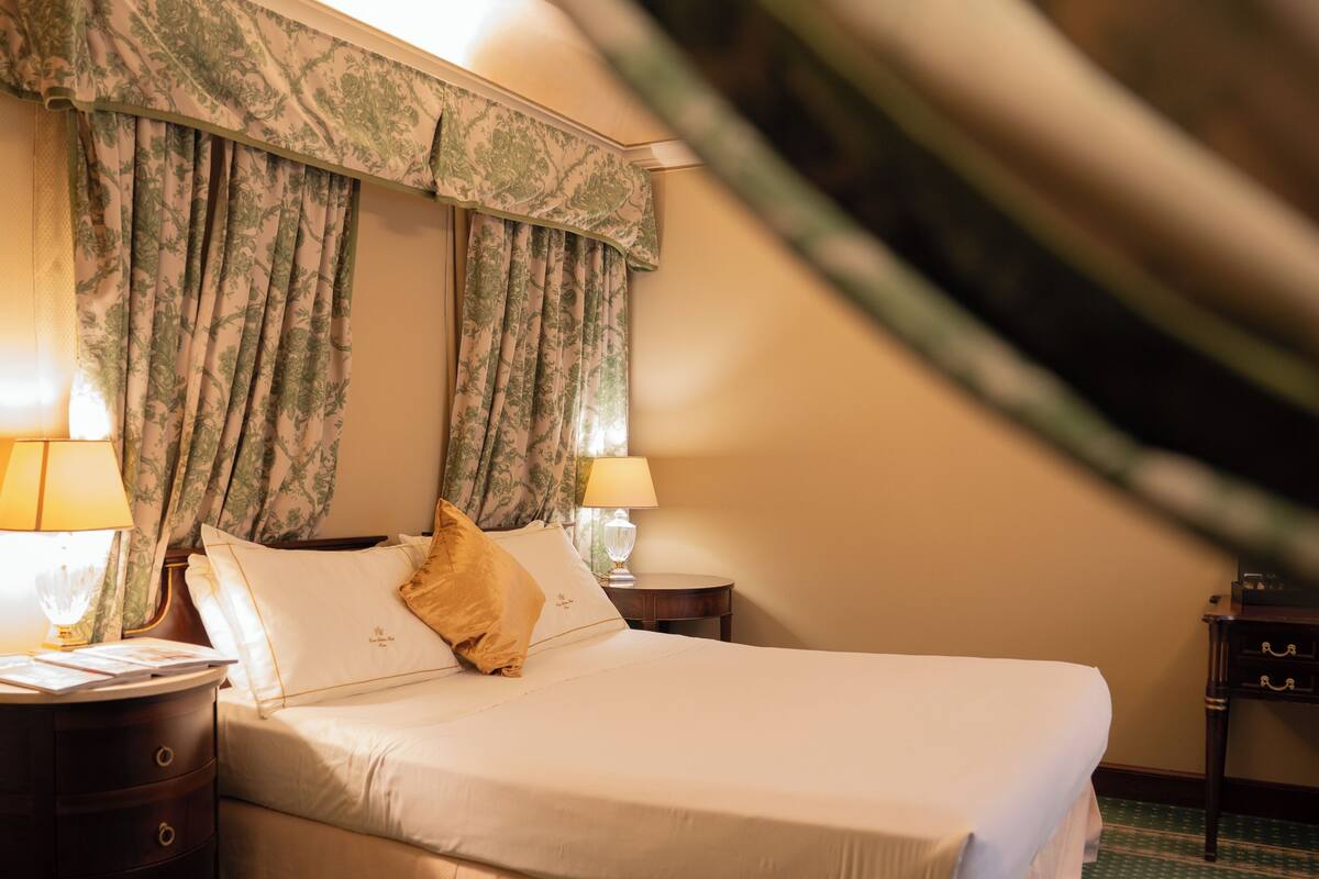 Hotellihuone, jossa on sänky, kaksi yöpöytälamppua, yöpöytä ja ikkuna, jossa on verhot.