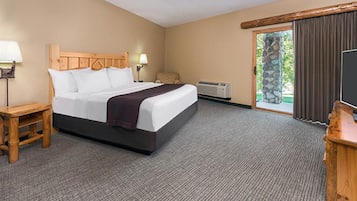 A hotel room with a large bed, a wooden headboard, a desk with a lamp, a television, and a view of the outdoors through a glass door.