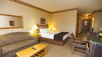 Habitación de hotel con sofá, cama con cabecera marrón, mesa de centro de madera, mesa de comedor con sillas y lámpara a cada lado de la cama.
