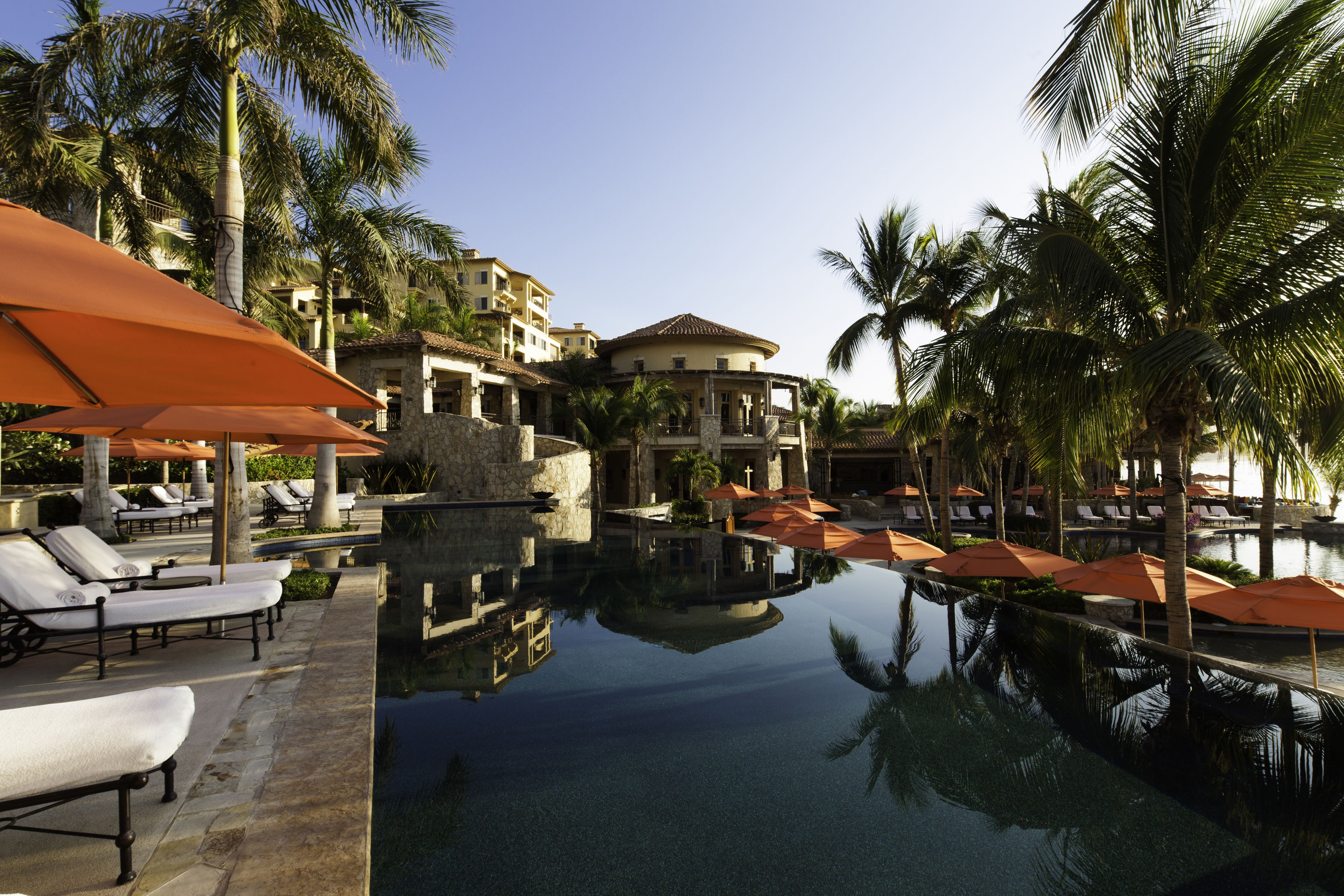 A resort pool area with lounge chairs and umbrellas, surrounded by palm trees and a building with a tiled roof.
