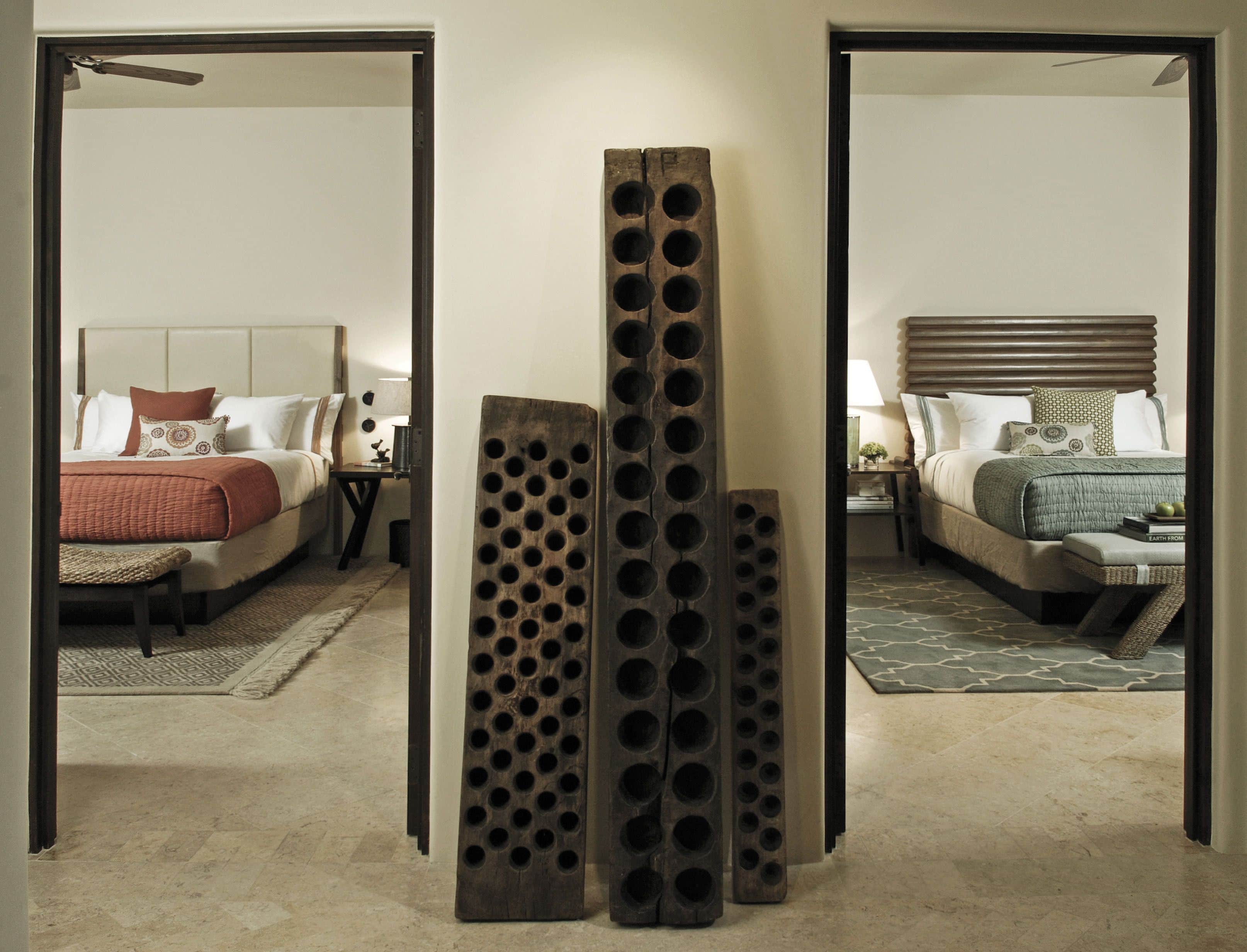 Ein Hotelzimmer mit einem großen Bett, einem Nachttisch und einem Sessel. Im Zentrum des Raumes befindet sich eine Holzskulptur.