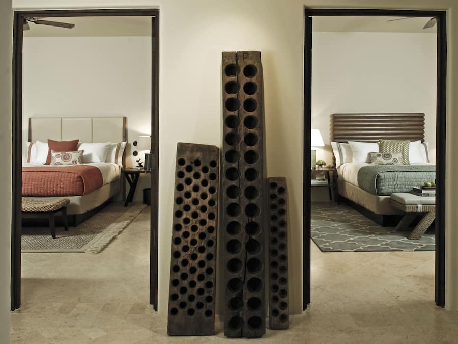 Ein Hotelzimmer mit einem großen Bett, einem Nachttisch und einem Sessel. Im Zentrum des Raumes befindet sich eine Holzskulptur.