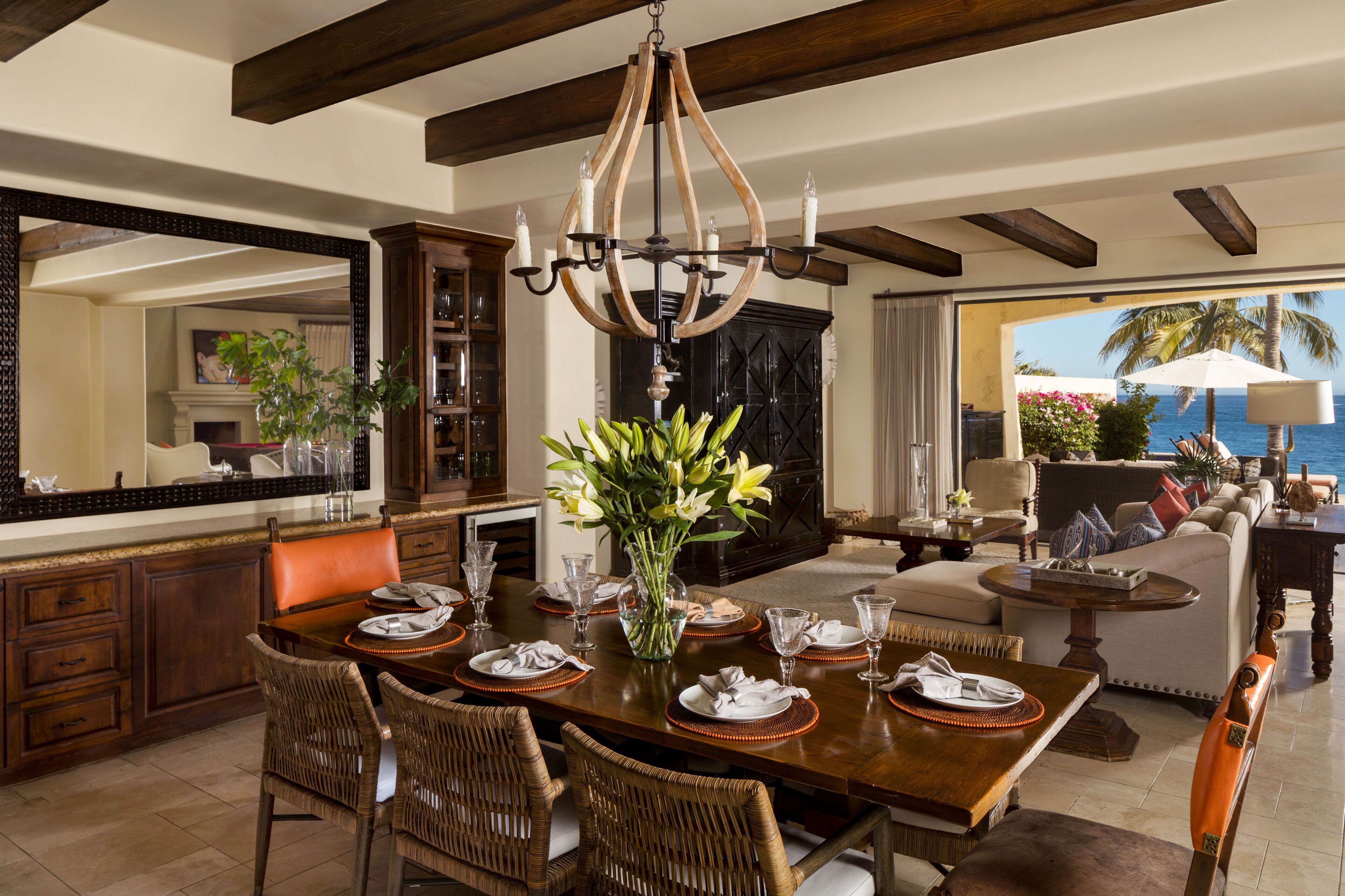 Un coin repas avec une grande table en bois, dressée pour six personnes, entourée de chaises en osier. Un lustre est suspendu au-dessus de la table, et un grand miroir reflète la pièce.