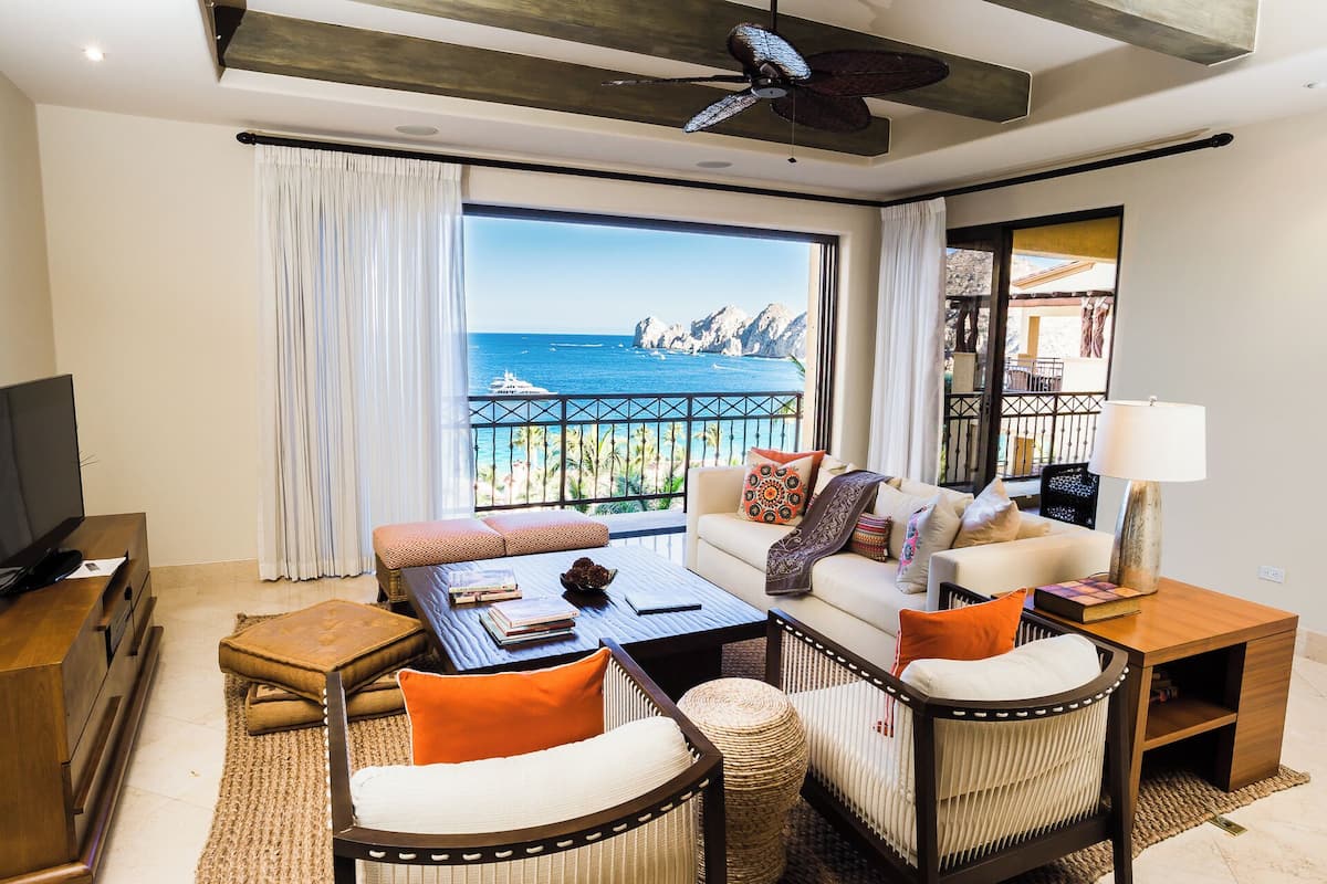 A modern living room with a balcony overlooking a coastal view, furnished with a sofa, chairs, a coffee table, and a TV.