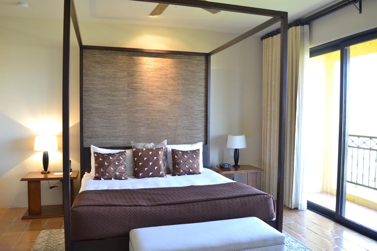 A modern hotel room with a large bed, a nightstand, a lamp, and a view of the outside balcony.