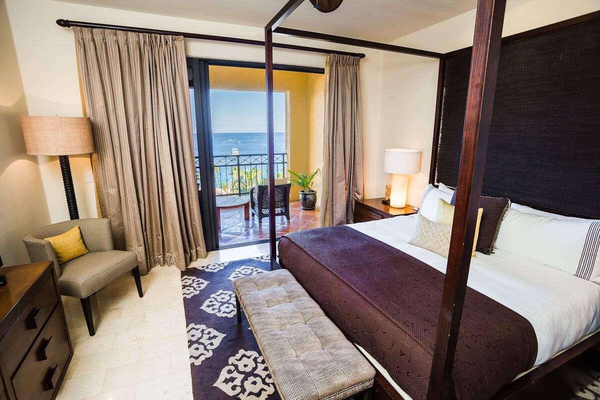 A four-poster bed with a view of the sea, a sitting area with a chair and a table, and a balcony with a view.
