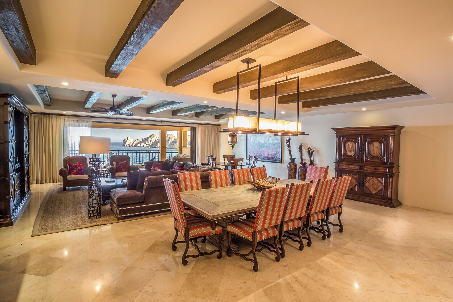 A spacious dining area with a large wooden table and striped chairs, a chandelier, and a view of the sea through large windows.