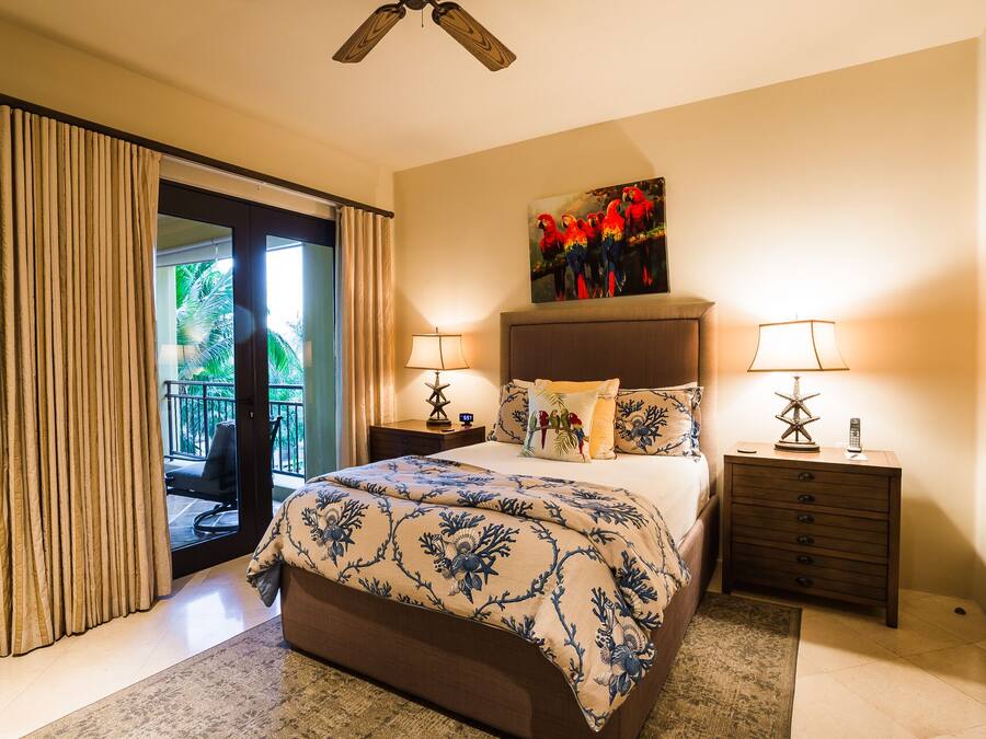 Quarto com uma cama grande, duas mesas de cabeceira com luminárias, um quadro de araras e vista para uma varanda com cortinas.