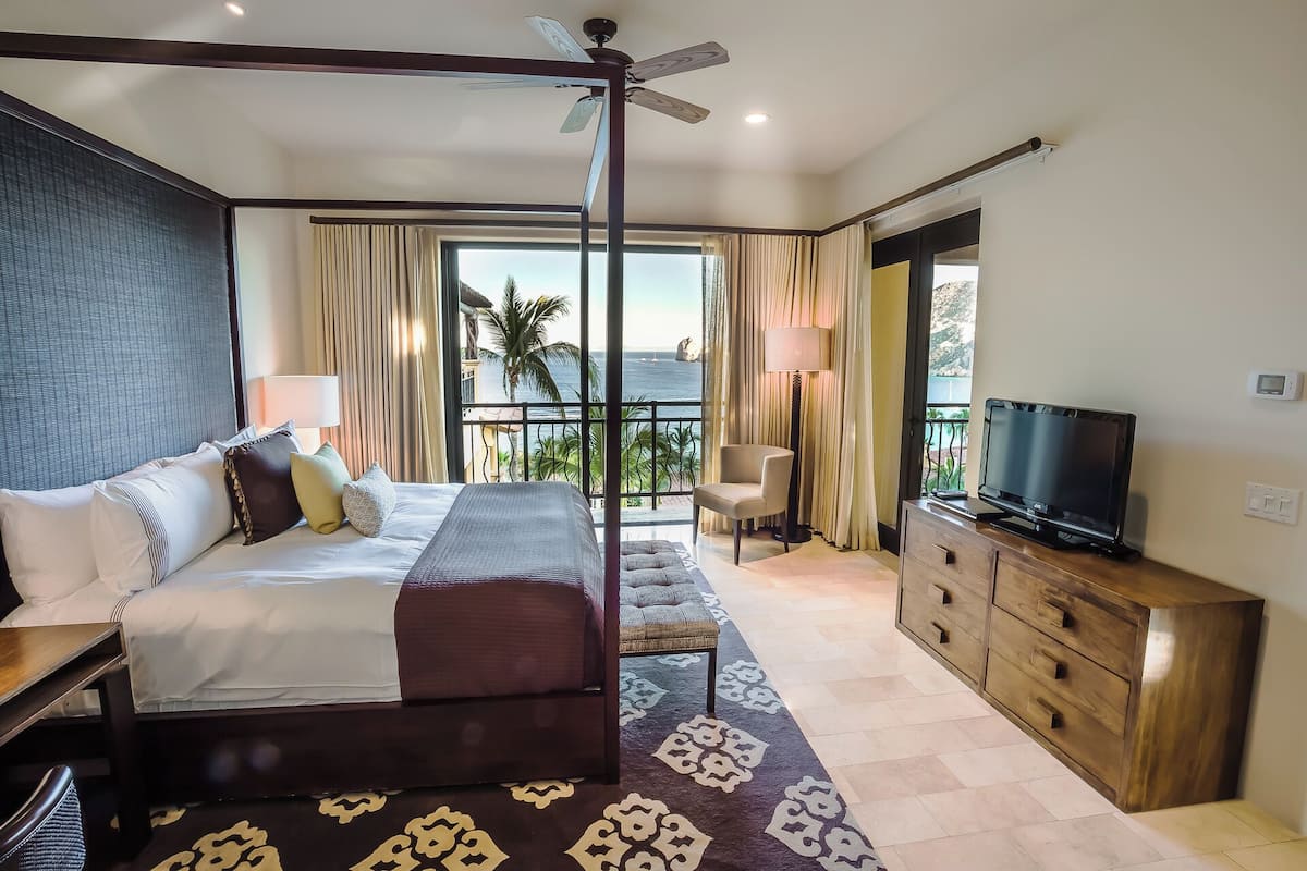 A spacious bedroom with a four-poster bed, a television, a wooden dresser, and a view of the ocean.