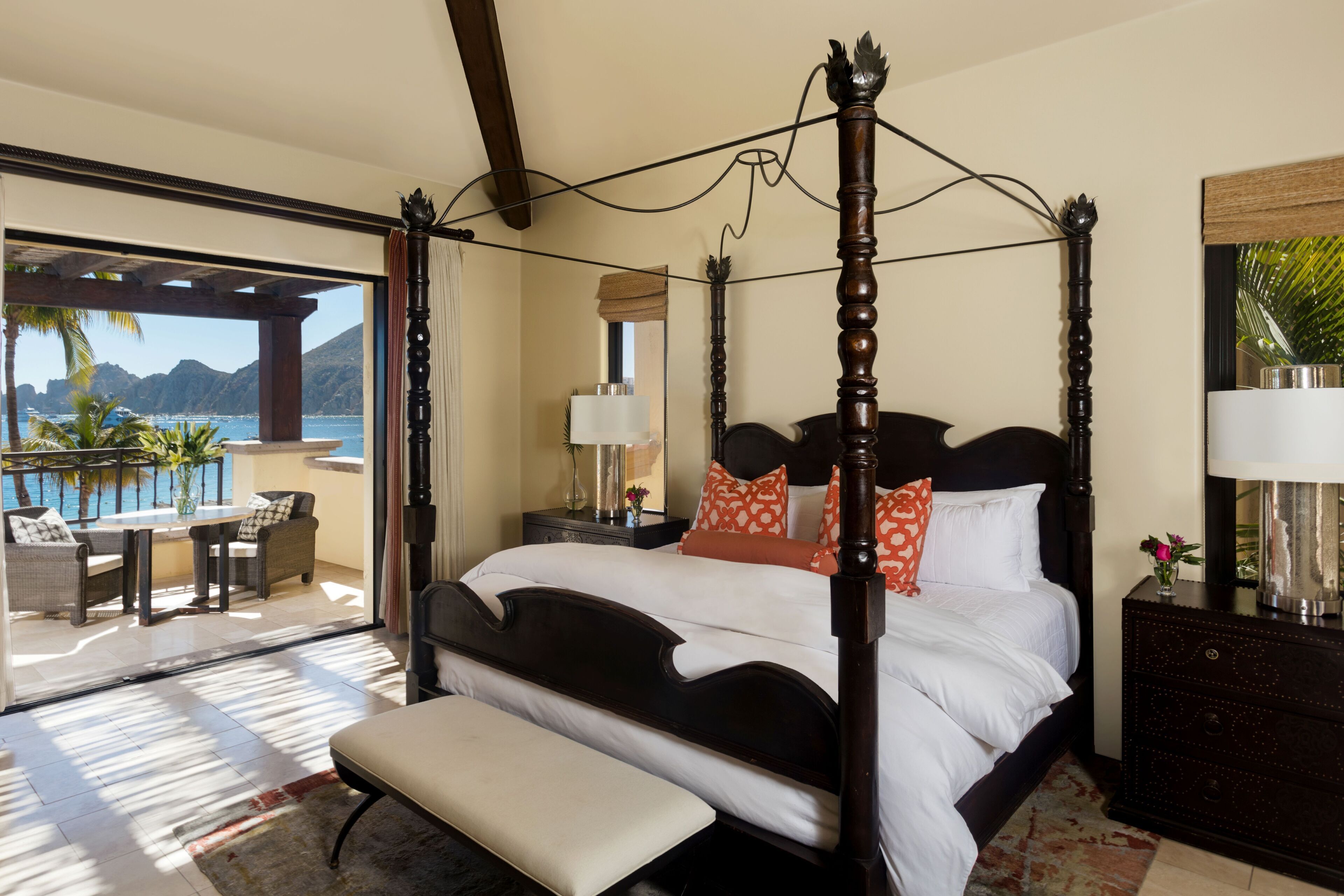 A four-poster bed with white linens, a bench, and a nightstand with a lamp.
