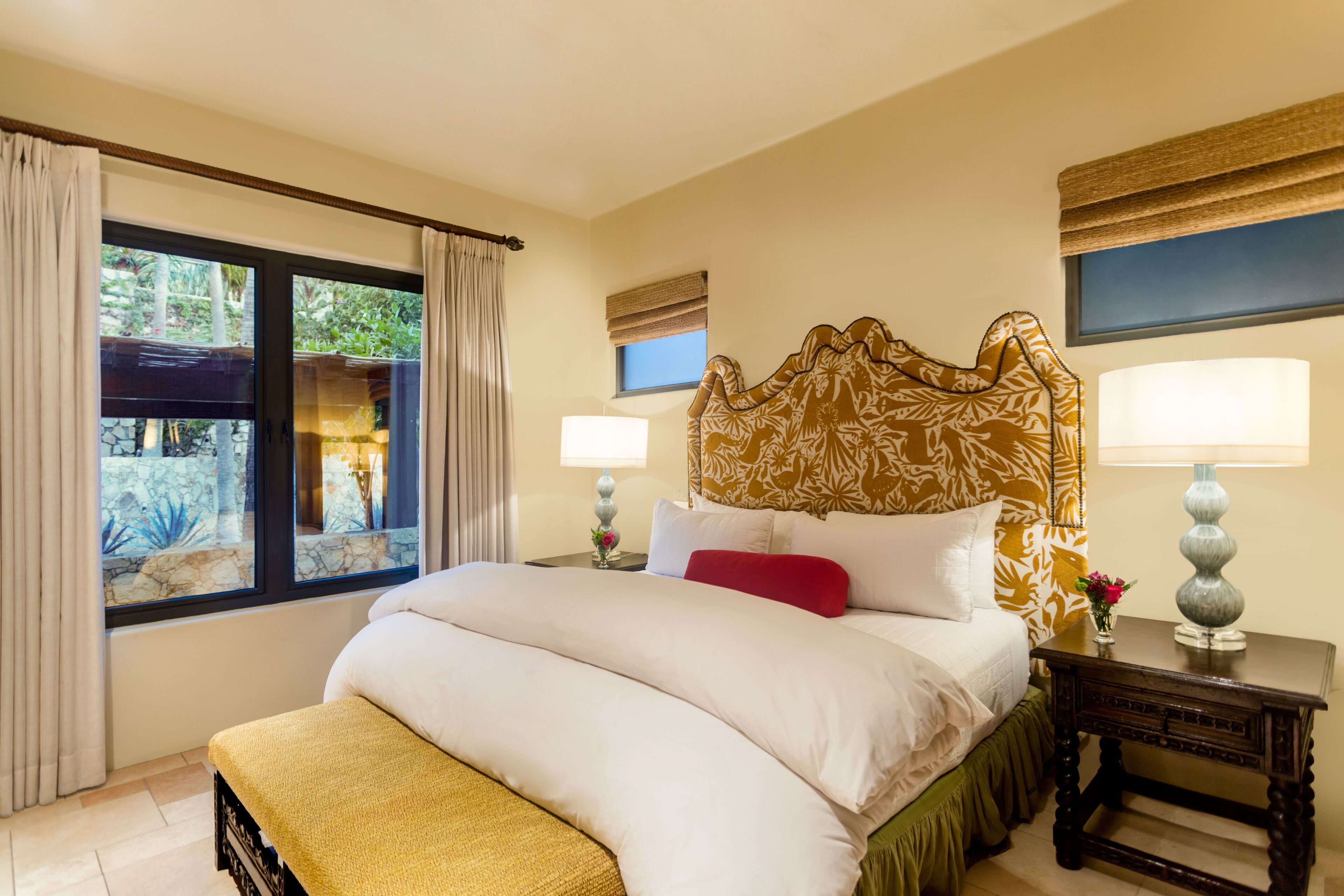 A bedroom with a large bed, two bedside tables with lamps, a window with curtains, and a view of a garden.
