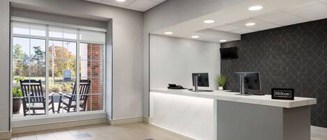 A modern hotel reception area with a large window, a reception desk, and a seating area with chairs.