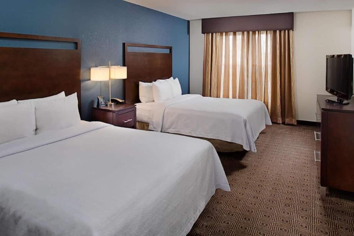 Habitación de hotel con dos camas, televisión, escritorio con lámpara y alfombra estampada.
