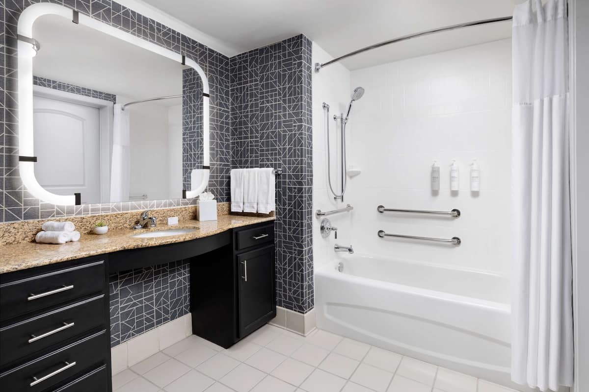 Un baño moderno con un espejo grande, encimera de granito, armarios negros, una bañera blanca y una zona de ducha.