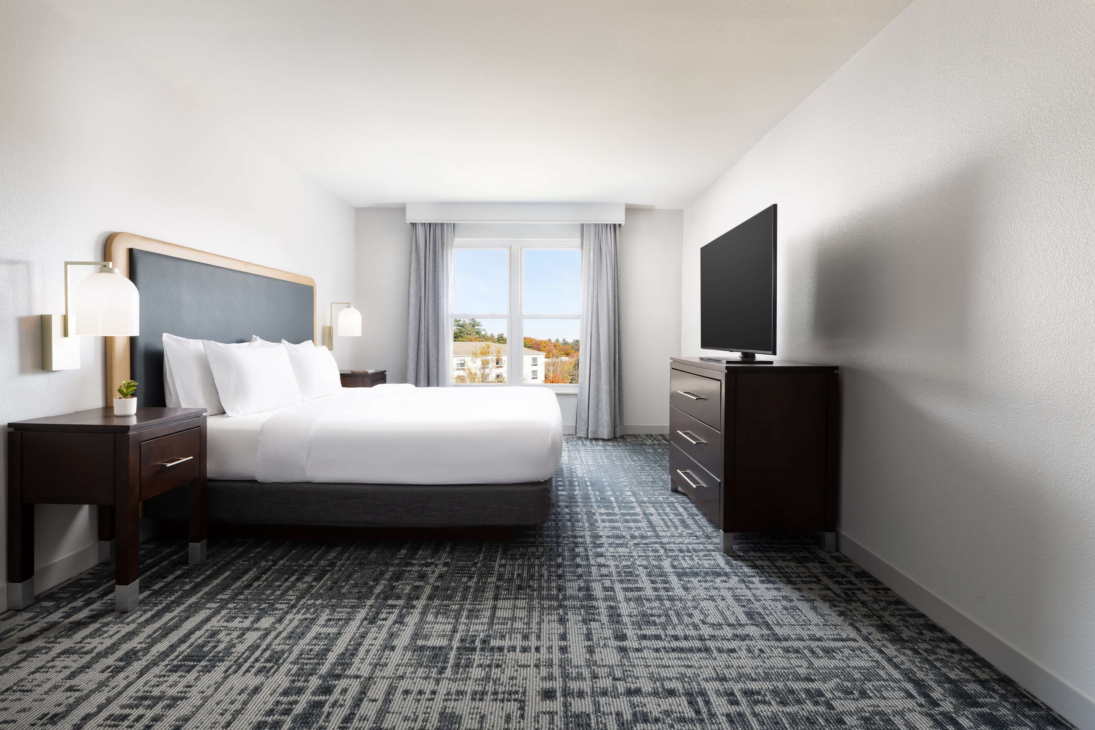A hotel room with a large bed, a TV, a desk with a lamp, and a view of buildings through the window.
